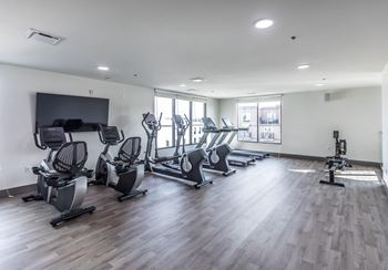 a gym with cardio equipment on a wooden floor and windows